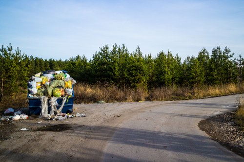 A blue waste disposal bin situated on a gravel roadside area, overflowing with a variety of discarded rubbish including plastic bags filled with mixed waste, cardboard boxes, and other household refuse, with loose debris and trash scattered on the ground nearby. The bin is positioned close to a bend in the road, with a backdrop of dense green trees and hedgerows under a partly cloudy sky. The scene appears to be an outdoor location, possibly in a rural or suburban area near Finchley, illustrating the type of rubbish accumulation that Finchley House Clearance services might help manage and remove from residential or green spaces in the region.