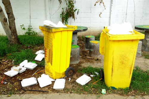 In an outdoor urban environment, two large yellow plastic waste bins with black wheels and closed lids are positioned side by side on a paved surface, surrounded by scattered white foam takeout containers and a few small pieces of litter. The background features a white painted brick wall with some green foliage and a tree trunk visible on the left. The scene suggests improper disposal of household rubbish, with the scattered containers and waste on the ground, highlighting the need for professional rubbish removal services such as those provided by Finchley House Clearance in North London, especially in areas like Finchley or nearby postcode regions.