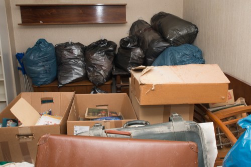 The image shows a cluttered indoor space, possibly a storage room or corner of a house, filled with multiple black and blue plastic rubbish bags stacked on and against a wooden bench or shelf, and several cardboard boxes containing miscellaneous items such as paper, books, and small objects. A large cardboard box with a torn lid is positioned at the front, partially obscuring other items behind it. To the right, there is a grey vacuum cleaner and an orange or reddish-brown leather or faux-leather piece of furniture or cushion. The background features a beige wall with a wooden shelf mounted above the bags. The overall scene suggests an area awaiting clearance or rubbish removal services, like those provided by Finchley House Clearance, to tidy up accumulated waste and unwanted belongings. The lighting appears to be natural or ambient, creating a neutral and professional atmosphere appropriate for a rubbish collection context within a residential or commercial property in the Finchley area.