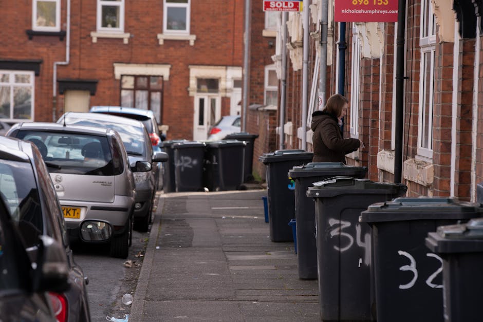 A row of black waste disposal bins lined up along the sidewalk of a residential street, with a woman standing next to one of the bins, opening or closing the lid. The bins are positioned in front of b