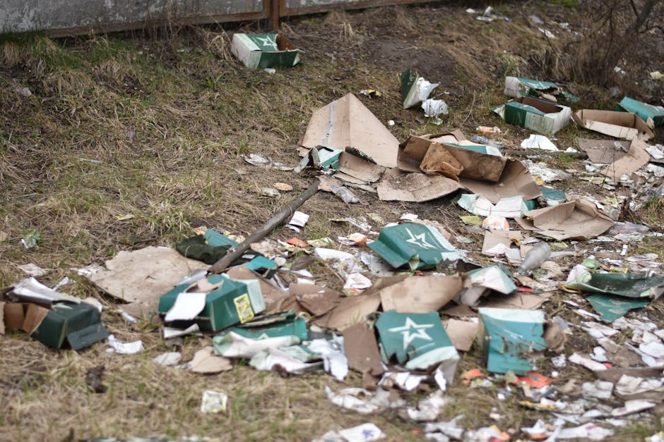 A collection of discarded waste materials piled on an asphalt surface in an outdoor area, including black plastic bin bags, a yellow plastic storage box, a worn car tire, and various miscellaneous rub